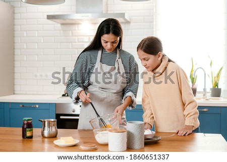 Focused beautiful mother and daughter smiling while making pancake dough in cozy kitchen