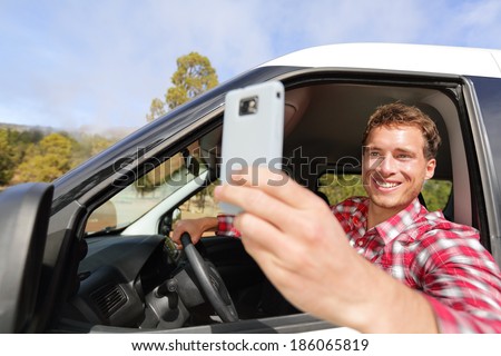 Driver taking photo with camera smartphone driving in car. Happy man taking picture with smart phone camera out window of car during travel road trip. Young Caucasian male model in his 20s.