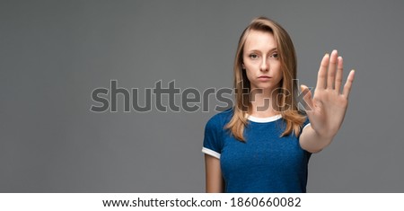 Serious girl with blonde straight hair in blue t shirt shows a stop with her hand, gesturing finish, showing symbol of rejection, not allow violence, body language. Copy space for your text Royalty-Free Stock Photo #1860660082