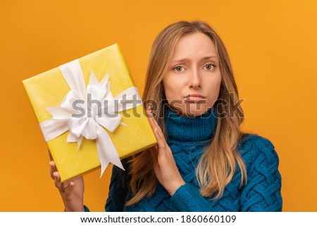 Portrait of a charming young woman in knitted blue sweater holding gift decorated with ribbon. Studio shot, yellow background. New Year, Women's Day, Birthday, Holiday concept