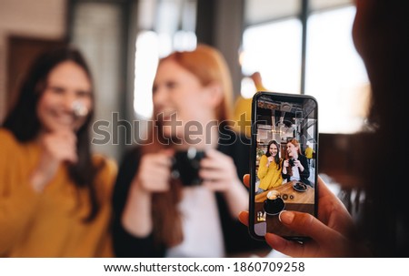 Woman sitting at a restaurant taking photograph of her friends having coffee. Woman shooting pictures with mobile phone of female friends at cafe.
