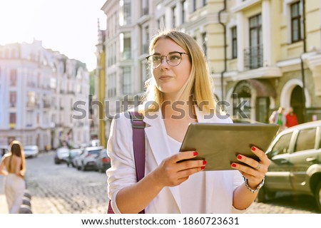 Young business woman in city using digital tablet. Beautiful smiling woman in white jacket with glasses looking at tablet monitor, urban style background