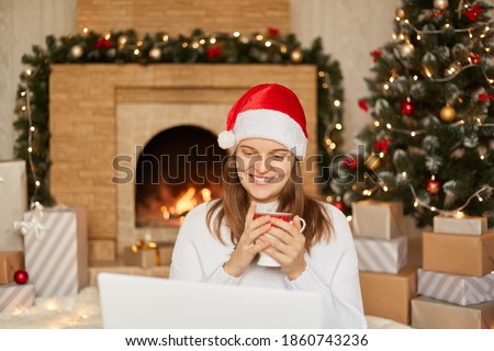 Photo of young lady sit in front of laptop, holding cup with hot tea, look at screen screen, wearing red santa hat and white sweater in decorated x-mas living room, congratulates somebody with holiday