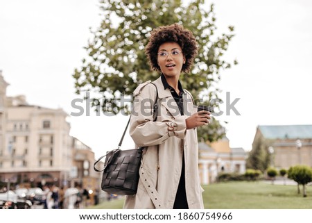 Charming young brunette curly woman in beige trench coat holds leathered black handbag and coffee glass outside.