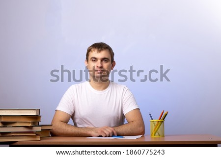 a young male student or teacher sits at a desk with books and pencils on the background of a gray wall. smile and look at the camera. space for text