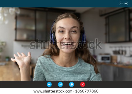Head shot sincere millennial happy young caucasian woman in headphones waving hand making hello gesture. Female greeting friends or family starting online video call meeting at home, feeling excited. Royalty-Free Stock Photo #1860797860