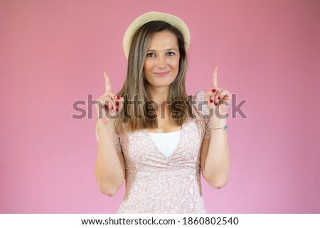 Smiling beautiful woman in pink dress and straw hat pointing up and looking at the camera over pink background