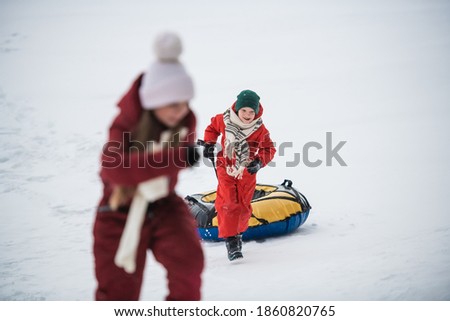 Winter walk of children in red overalls, a boy carries his tubing up the hill.