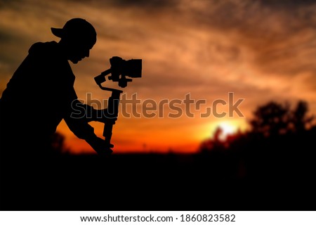 Silhouette of videographer is filming with cinema gimbal video dslr at sunset , professional video, videographer in events. Cinema lens on gimbal. Medium shot from right side. Film or cameraman school Royalty-Free Stock Photo #1860823582