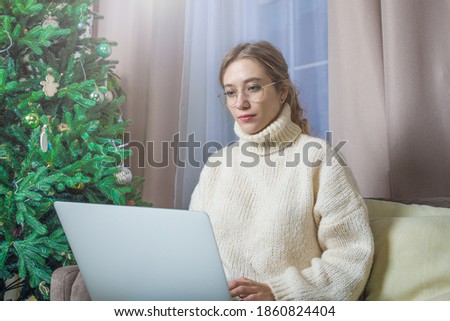 Young beautiful woman in glasses and trendy warm sweater checking e-mail on laptop computer while sitting in home near Christmas tree during the winter break celebration