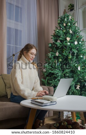 Gorgeous female student having online education via laptop computer while sitting in home near Christmas tree during winter holidays. Woman writer typing article via netbook during December vacation 