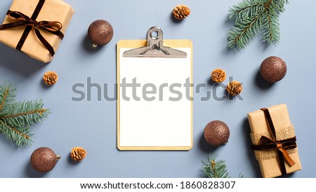 Festive workspace table with blank paper clipboard, gift boxes, fir branches, balls on pastel blue background. Christmas, winter holidays concept. Minimal, nordic style.