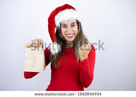 Young beautiful woman wearing a Santa hat over white background holding a paper bag very happy and excited making winner gesture with raised arms, smiling and screaming for success