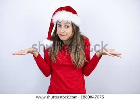 Young beautiful woman wearing a Santa hat over white background clueless and confused expression with arms and hands raised