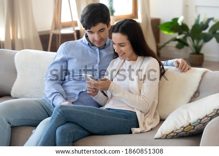 Online together. Happy loving young couple sitting on couch browsing internet or social networks using cellphone. Family making video call, chatting with friends, watching cute photos on gadget screen
