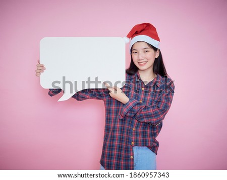 Cute teenage gir in santa hat pointing finger on blank poster while standing on pink background.
