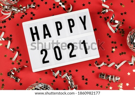 white lightbox with words happy 2021, disco ball, ribbon and holiday confetti on red background. top view. Happy New Year 2021 concept. 