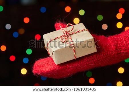 A child in a mitten holds a Christmas or New Year gift box against a background of colorful lights. Close-up, selective focus