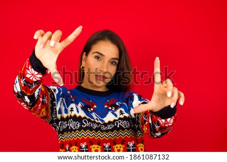 Portrait of smiling Young beautiful Hispanic woman wearing winter sweater against red wall looking at camera and gesturing finger frame. Creativity and photography concept.