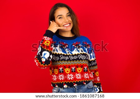 Young beautiful Hispanic woman wearing winter sweater against red wall imitates telephone conversation, makes phone call gesture with hands, has confident expression. Call me!