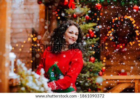Attractive brunette woman in red Christmas sweater posing on the open wooden terrace with Christmas decoration and snowing.