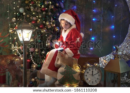 little Santa Claus holds a magic lantern and a sack  of gifts and looks at his glowing palm. Christmas tree decorated with balloons on a dark background with sparkling lights and magic garlands