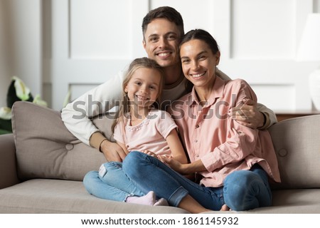 Loving young man hug his beloved wife and little daughter sit on couch in living room, happy people smiling looking at camera posing for photo picture. Exemplary family portrait, love and bond concept Royalty-Free Stock Photo #1861145932