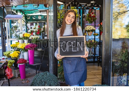 Smiling young florist in aorn holding open sign while standing in doorway of flower shop. Opening a shop. Happy florist standing in the doorway of her flower shop with an open sign.