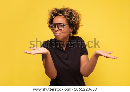 Young african woman with arms out, shrugging her shoulders, saying who cares, so what now. Studio shot on yellow wall. Royalty-Free Stock Photo #1861223659