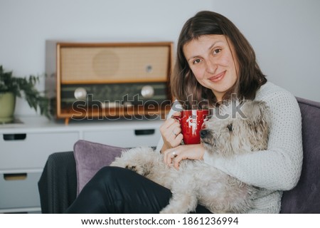 Happy woman with her dog enjoying morning coffee with her dog and listening to Christmas carols on an old-school radio. 