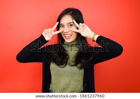 Portrait of Young beautiful asian women using black sweater making victory or peace sign using two hands with red isolated background