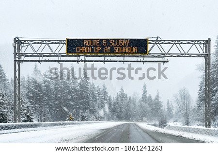 The road between tall spruce trees and mountain peaks in snowy weather. Road sign warning of dangerous road conditions. Coniferous snowy forest in the Rocky Mountains. British Columbia, Canada