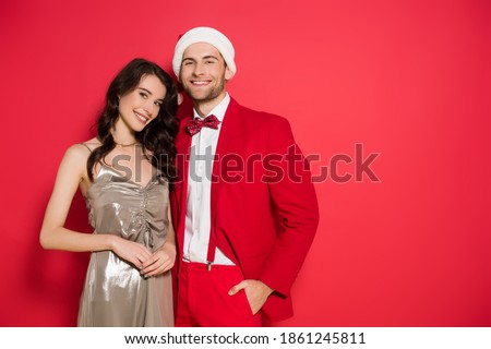 Smiling woman in dress looking at camera near stylish boyfriend in santa hat on red background