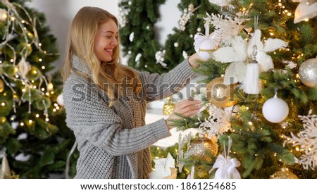 Happy young blond woman decorating christmas tree with ball at her home