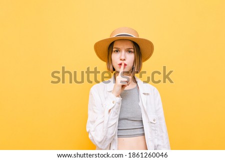 Attractive lady in light clothing and hat stands on a yellow background,looks in camera with a serious face and shows a gesture of silence with finger on lips.