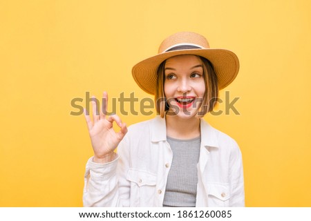 Closeup portrait of a funny woman in a hat and light clothing on a yellow background, looking to the side with a smile on her face and showing an OK gesture Copy space