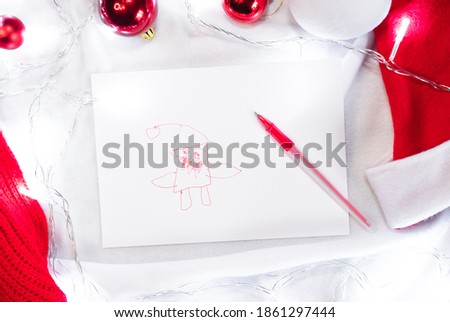 Creative children's drawing of Santa Claus on a white background. Cute Santa drawn by a child with a red pen and a white sheet of paper and around are red Christmas balls and lights garland.