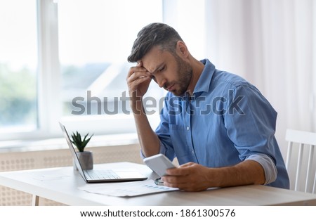 Worried male freelancer looking at smartphone screen while working at home office, received bad news, got scam message, thoughtful man touching forehead with concerned face expression, copy space Royalty-Free Stock Photo #1861300576
