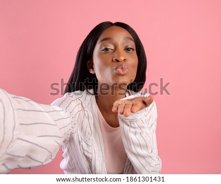 Attractive young black woman taking selfie, blowing air kiss on pink studio background. Lovely African American lady posing to mobile camera and making photo of herself