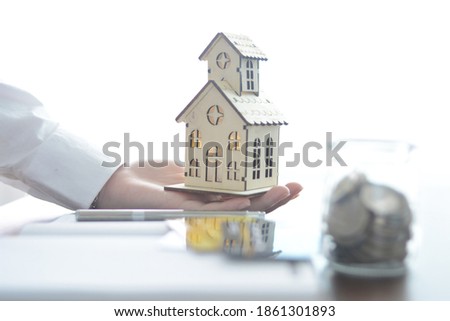 A wooden model house placed on a businessman's hand with a blurry image, a credit card and a glass jar in front of the photo.
