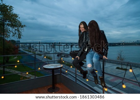 Two young brunette women with long hair posing in evening at sunset on embankment sitting on railing of summer roof of a cafe with garlands and bulbs against the background of the river and the bridge