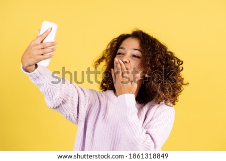 Charming young afro american woman making online video call on modern smartphone, talking with friends or family, sending air kiss, standing on yellow copy space background