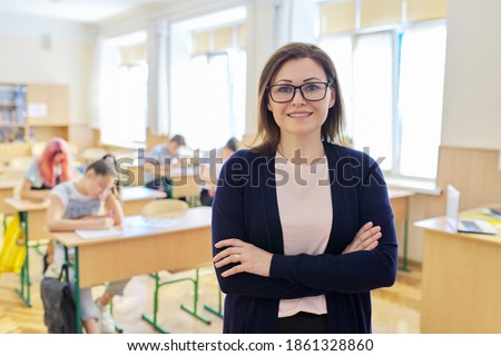 Portrait of female teacher at school in class, smiling middle aged woman wearing glasses with folded hands looking at camera, classroom with teenage students at desk background