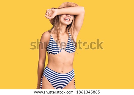 Young beautiful blonde woman wearing bikini and hat covering eyes with arm smiling cheerful and funny. blind concept. 