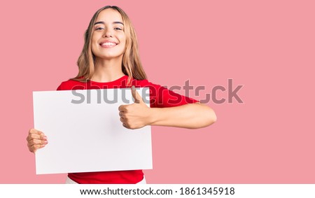 Young beautiful blonde woman holding blank empty banner smiling happy and positive, thumb up doing excellent and approval sign 