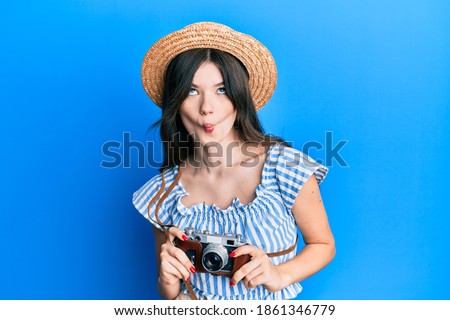 Young beautiful caucasian girl holding vintage camera making fish face with mouth and squinting eyes, crazy and comical. 