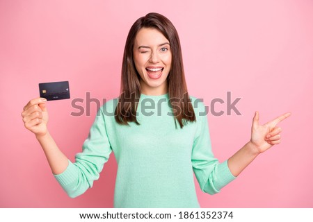 Portrait of positive funky brunette young woman point empty space wink promote credit card isolated on pink color background