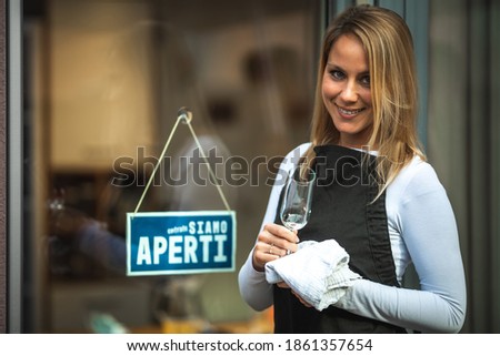 Young smiling Caucasian woman with a black apron, wiping a glass, in front of the door with a sign in Italian ''we are open''