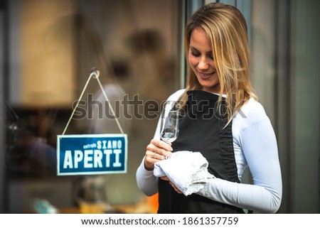 Young Caucasian woman with a black apron, wiping a glass, in front of the door with a sign in Italian ''we are open''