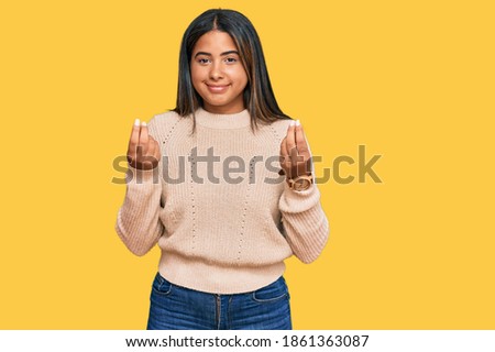 Young latin girl wearing wool winter sweater doing money gesture with hands, asking for salary payment, millionaire business 
